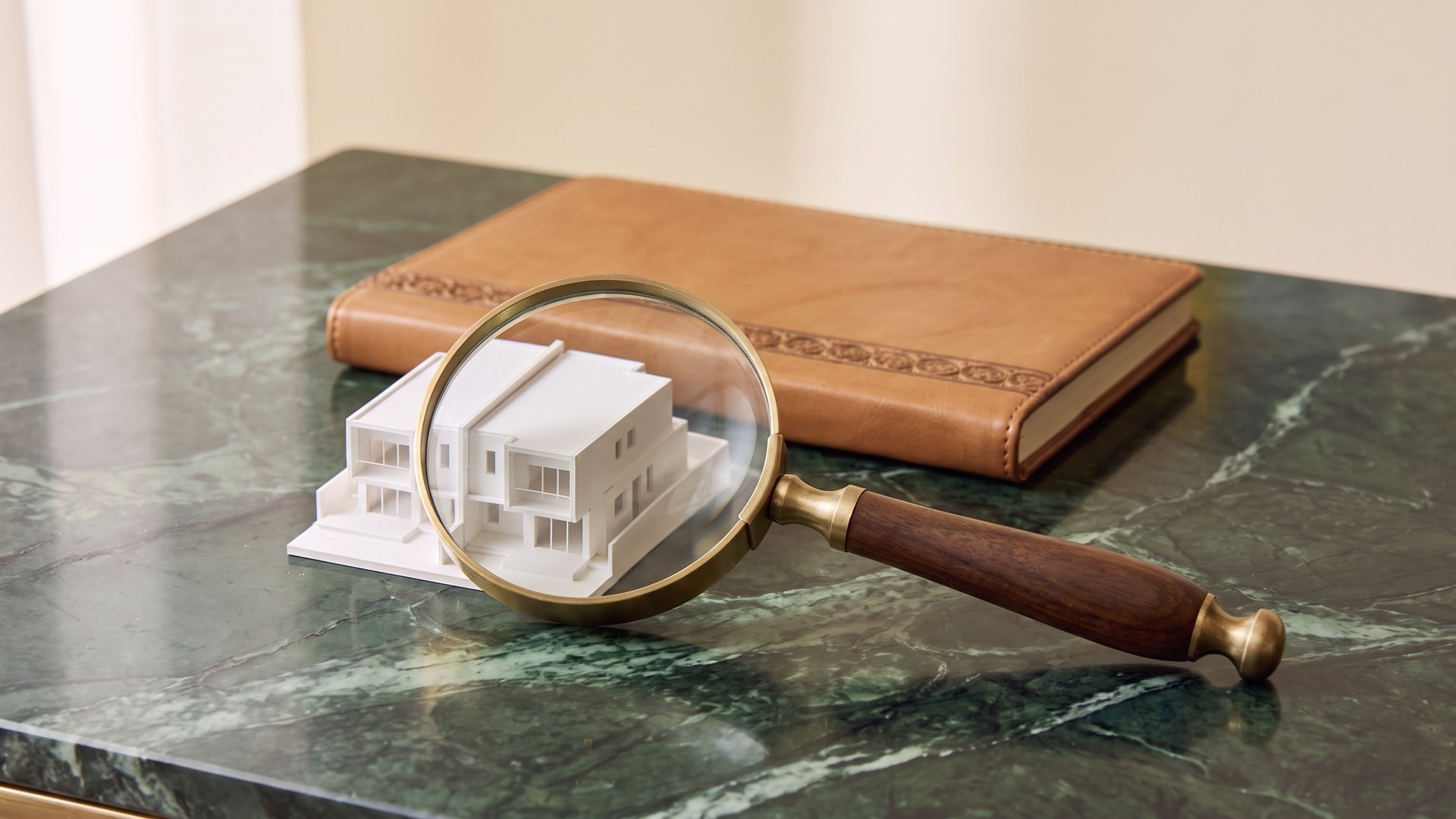 A brass magnifying glass with a dark wooden handle leans in front of a small white two-storey model house on a dark green veined marble desk, magnifying it, with a tan leather notebook with an embossed band behind.